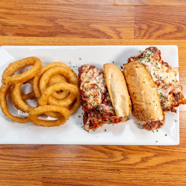 A white rectangular plate holds golden onion rings and two meatball subs with melted cheese.